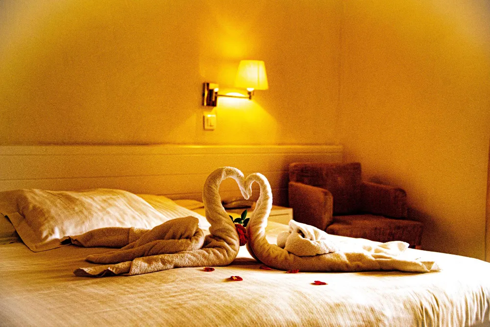 Lit double d’hôtel décoré de serviettes pliées en forme de cygnes formant un cœur, avec pétales de rose pour une ambiance romantique.